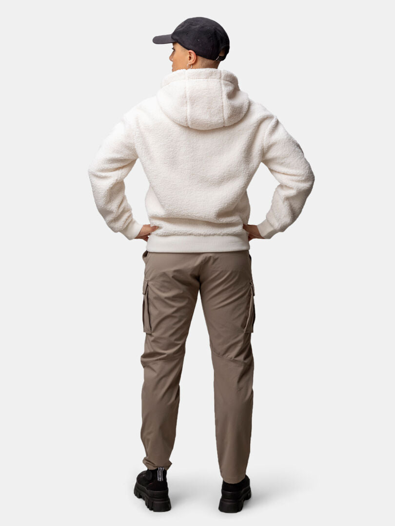 Henkilö takaapäin, kädet lanteilla, päällään Halti Lykky Fleecetakki Naisten, khakinväriset rahtitaskuhousut, mustat kengät ja lippis.