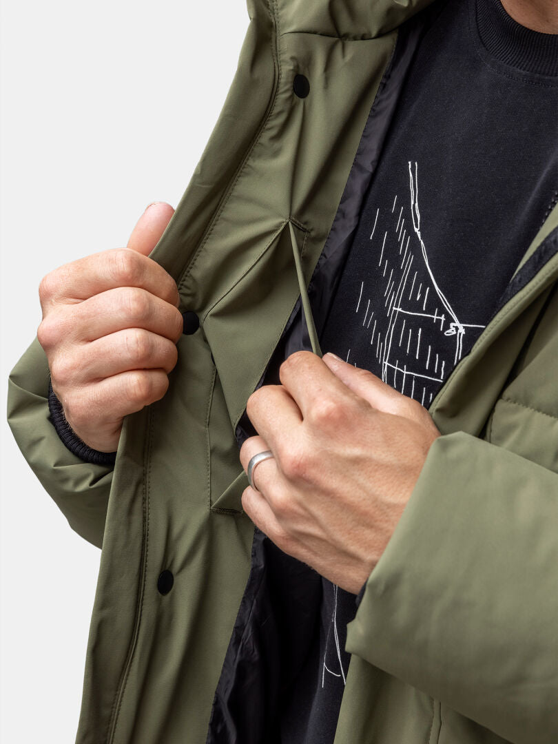 Henkilö säätää Halti Brahe Puffer Talvitakki Miesten sisällä olevaa kiristysnyöriä, kun hänellä on yllään musta graafinen paita.