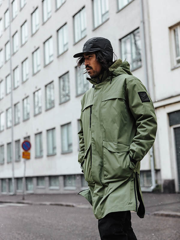 Halti Tokoi II DX Parkatakki -miehiin ja mustaan lippikseen pukeutunut henkilö kävelee valkoisen kerrostalon ohi kaupungissa.