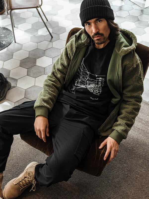 Halti Lykky Fleecetakki Miesten ja mustaan pipoon pukeutunut mies istuu ruskealla tuolilla kuusikulmaisella kaakeloidulla lattialla.