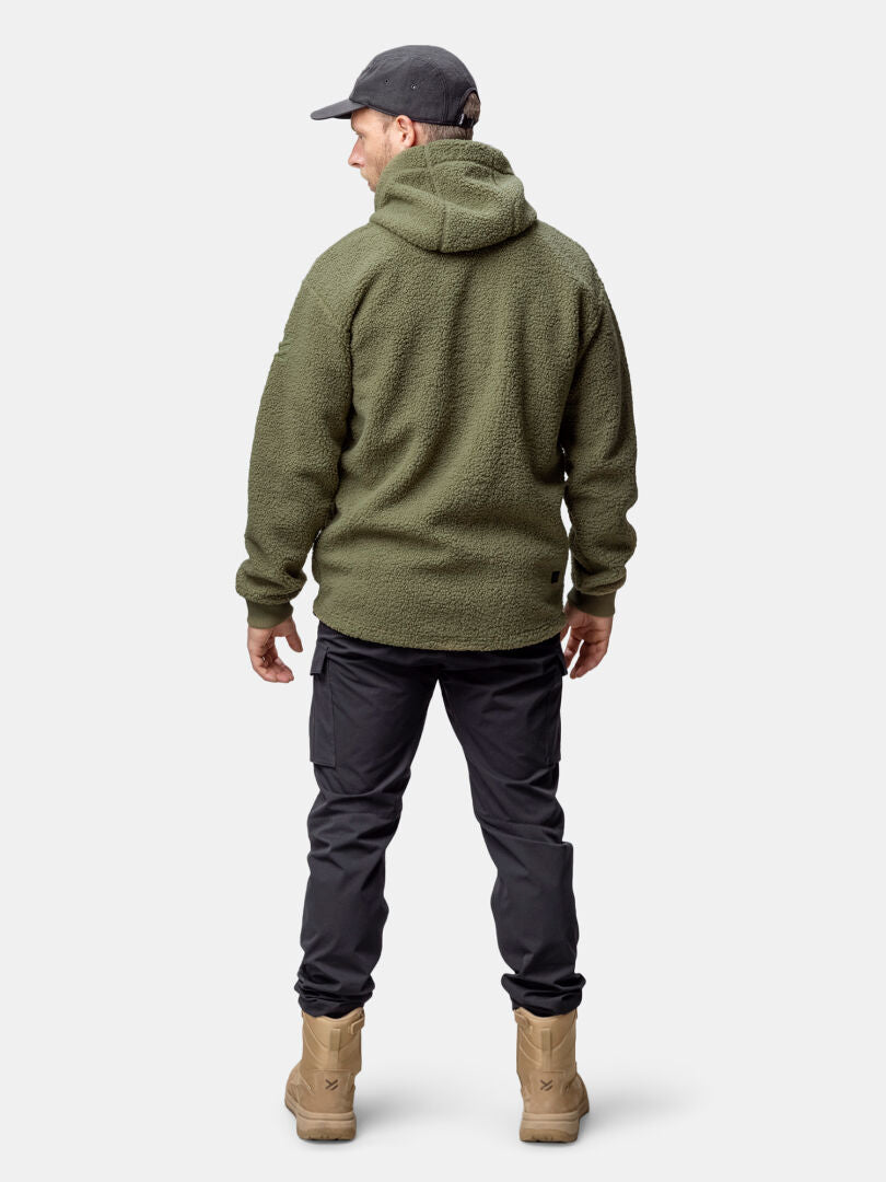 Halti Lykky Fleecetakki Miesten oliivinväriseen huppariin, mustiin housuihin, ruskehtaviin saappaisiin ja tummaan lippikseen pukeutunut mies katsoo kamerasta poispäin.