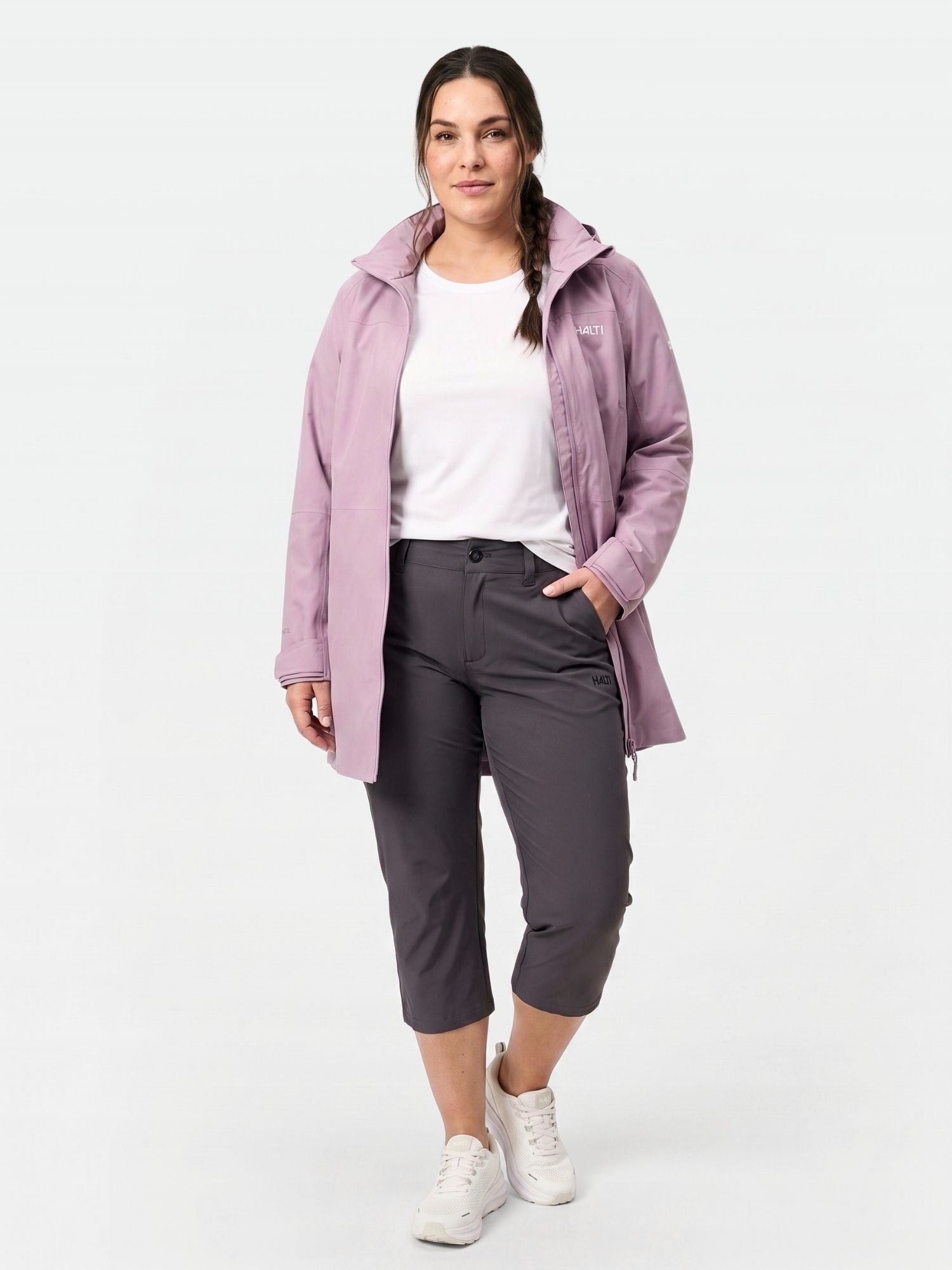 Naisella on Halti Drive X-stretch Caprit Naisten Plus, vaaleanvioletti takki, valkoinen t-paita ja valkoiset lenkkarit yksivärisellä pohjalla.