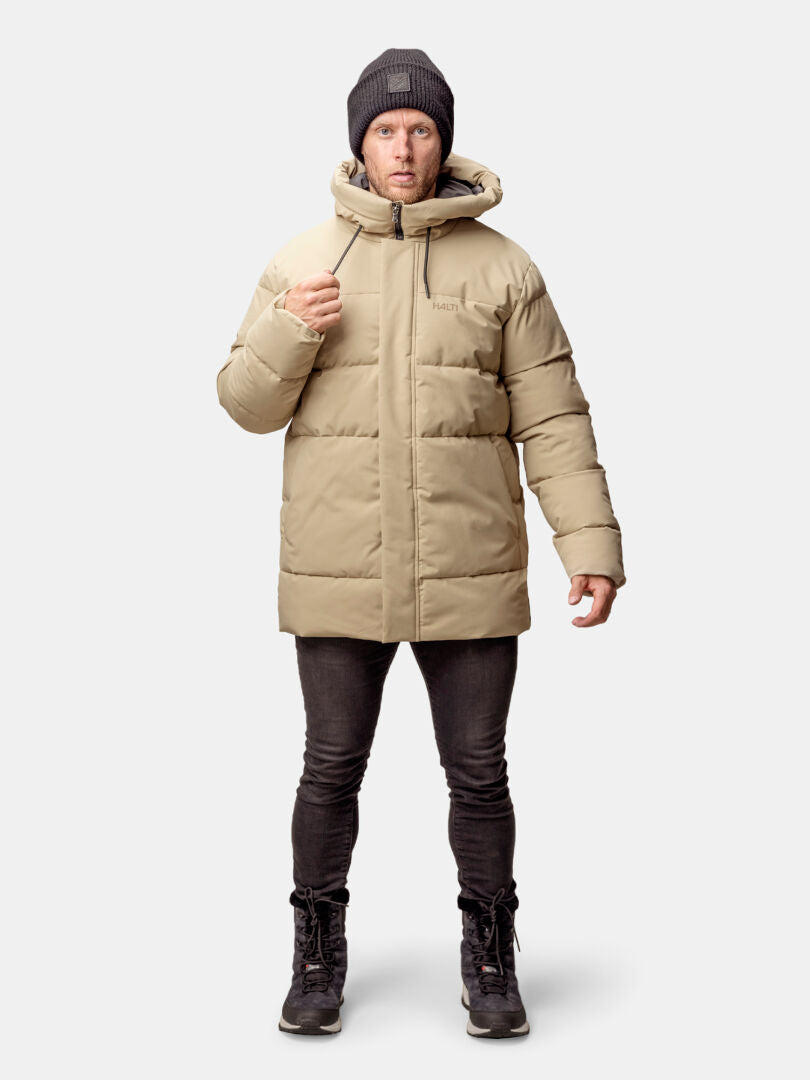 Henkilöllä on yllään beige Halti Sammal Topattu Parkatakki Miesten, musta pipo, mustat farkut ja saappaat yksiväristä taustaa vasten.