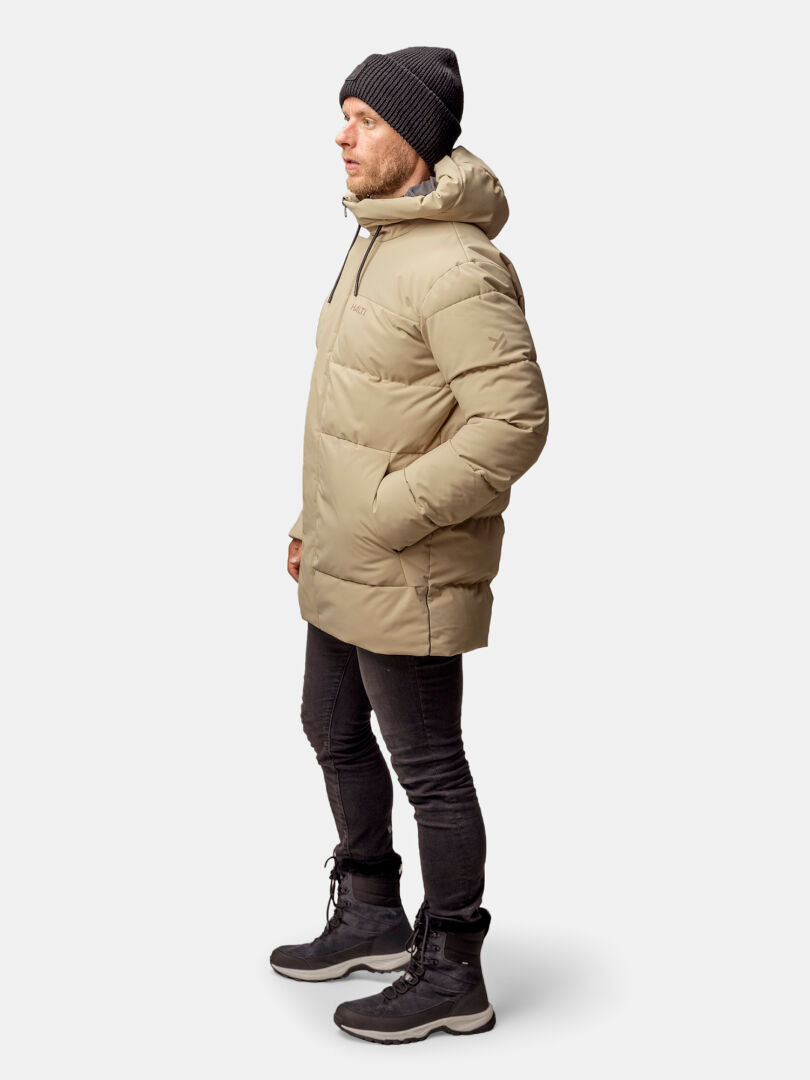 Profiilissa seisoo beigeen Halti Sammal Topattu Parkatakki Miesten, mustaan pipoon, housuihin ja saappaisiin pukeutunut mies.