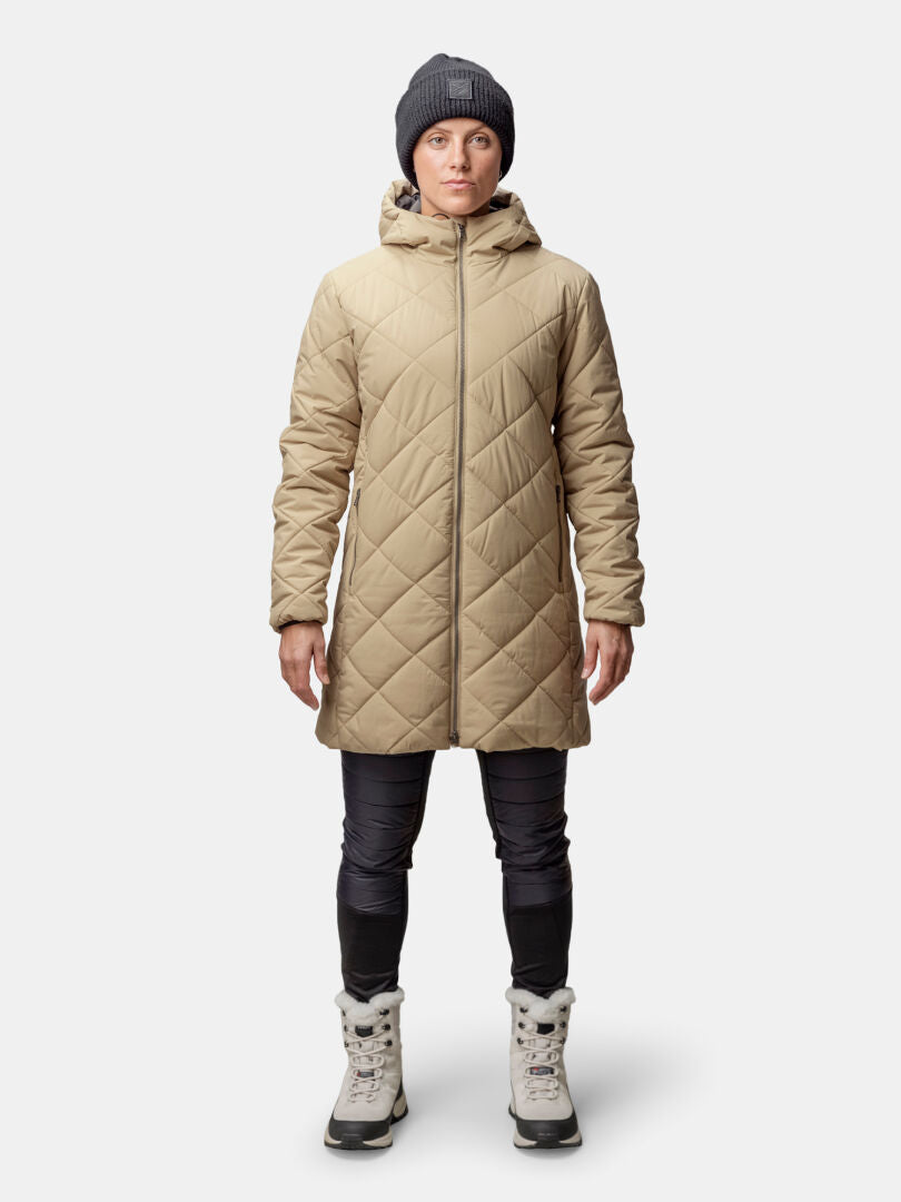 Henkilöllä on yllään Halti Haapa Tikkitakki Naisten takki, harmaa pipo, mustat housut ja vaaleanharmaat talvisaappaat tavallista taustaa vasten.