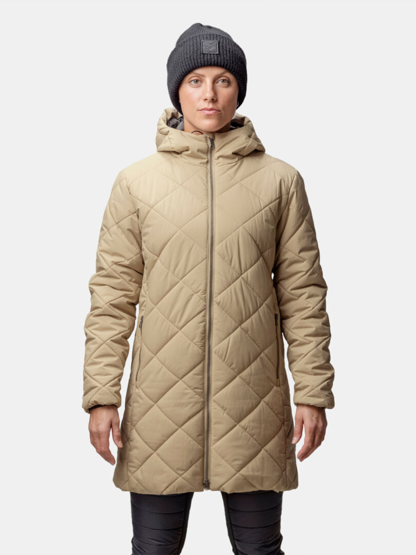 Henkilöllä on yllään beige Halti Haapa Tikkitakki Naisten, harmaa neuletakki ja tummat housut, ja hän seisoo tavallista taustaa vasten.