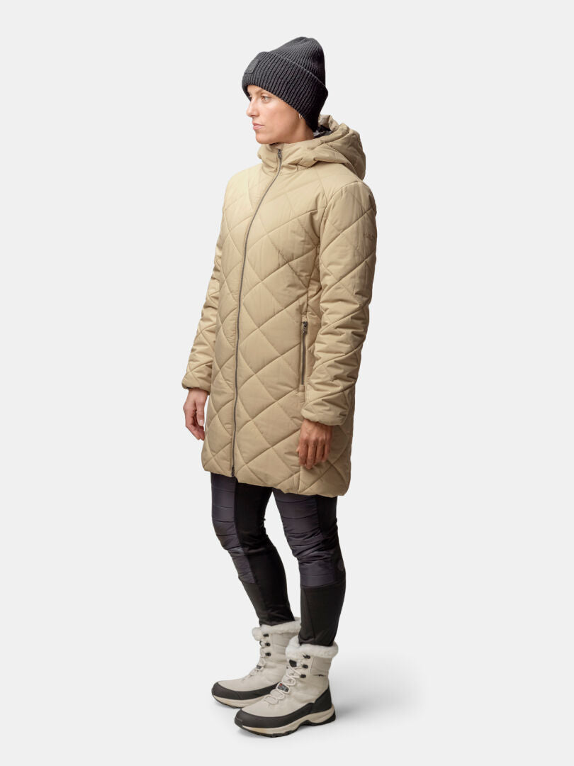 Henkilö, jolla on beige Halti Haapa Tikkitakki Naisten, mustat leggingsit, harmaat saappaat ja tumma pipo, seisoo tavallista taustaa vasten.