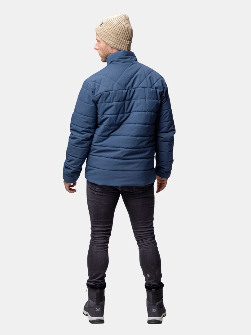 Mies, jolla on sininen Halti Haapa Kevyttoppatakki, mustat farkut, saappaat ja beige pipo, seisoo kasvot menosuuntaan vaalealla taustalla.