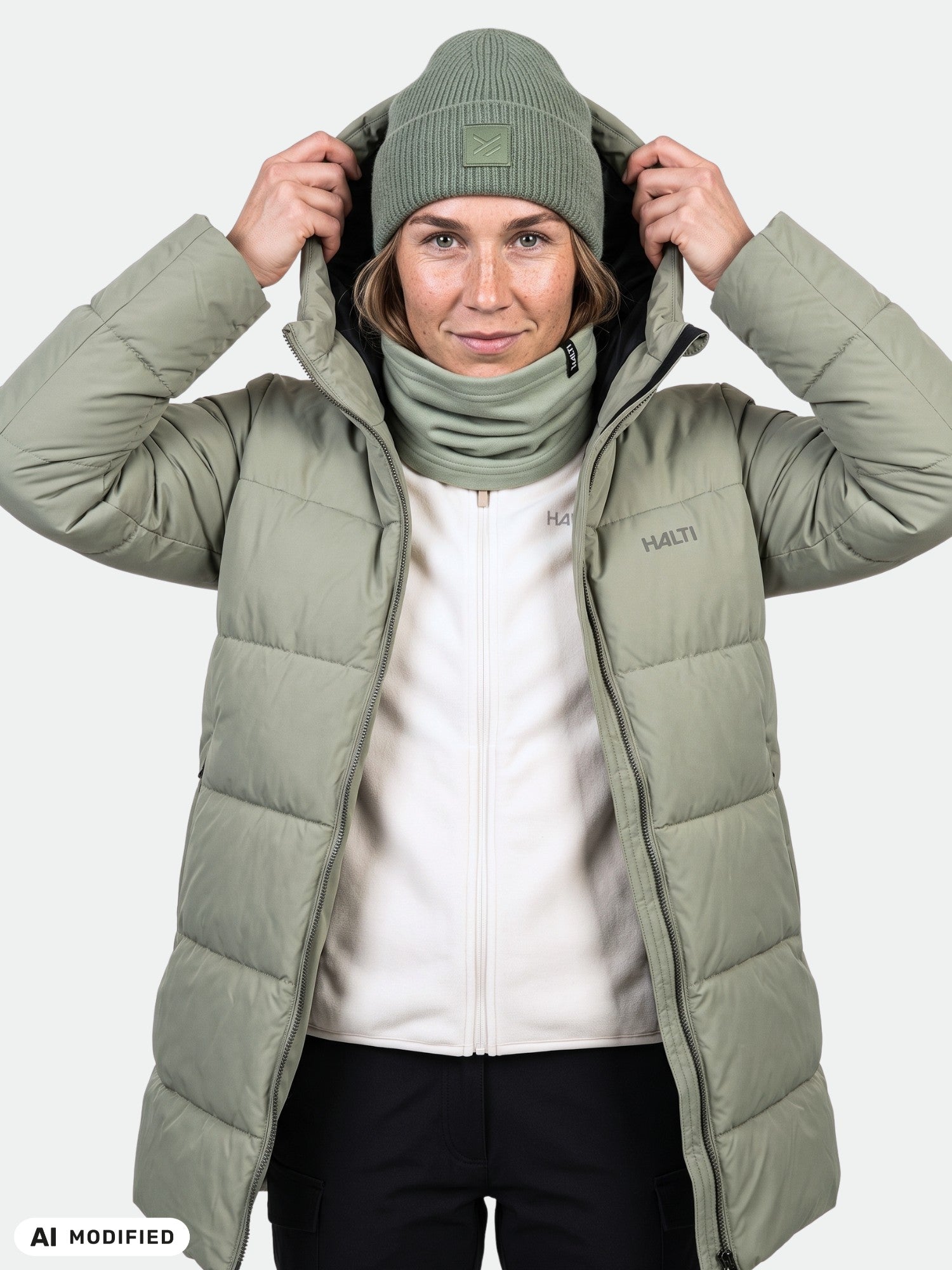 Nainen, jolla on yllään vaaleanvihreä Halti Leinikko Puffer Parka Naisten -takki, pipo ja kaulahuivi, säätää huppuaan niin, että se osoittaa eteenpäin.