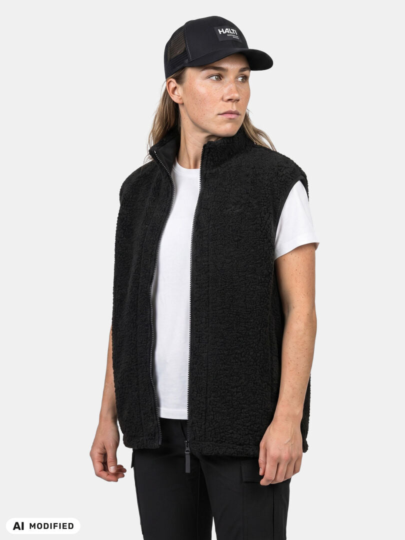 Joku, jolla on yllään Halti Lykky Fleece Liivi Unisex -liivi, valkoinen t-paita ja musta lippalakki, seisoo yksivärisen taustan edessä.