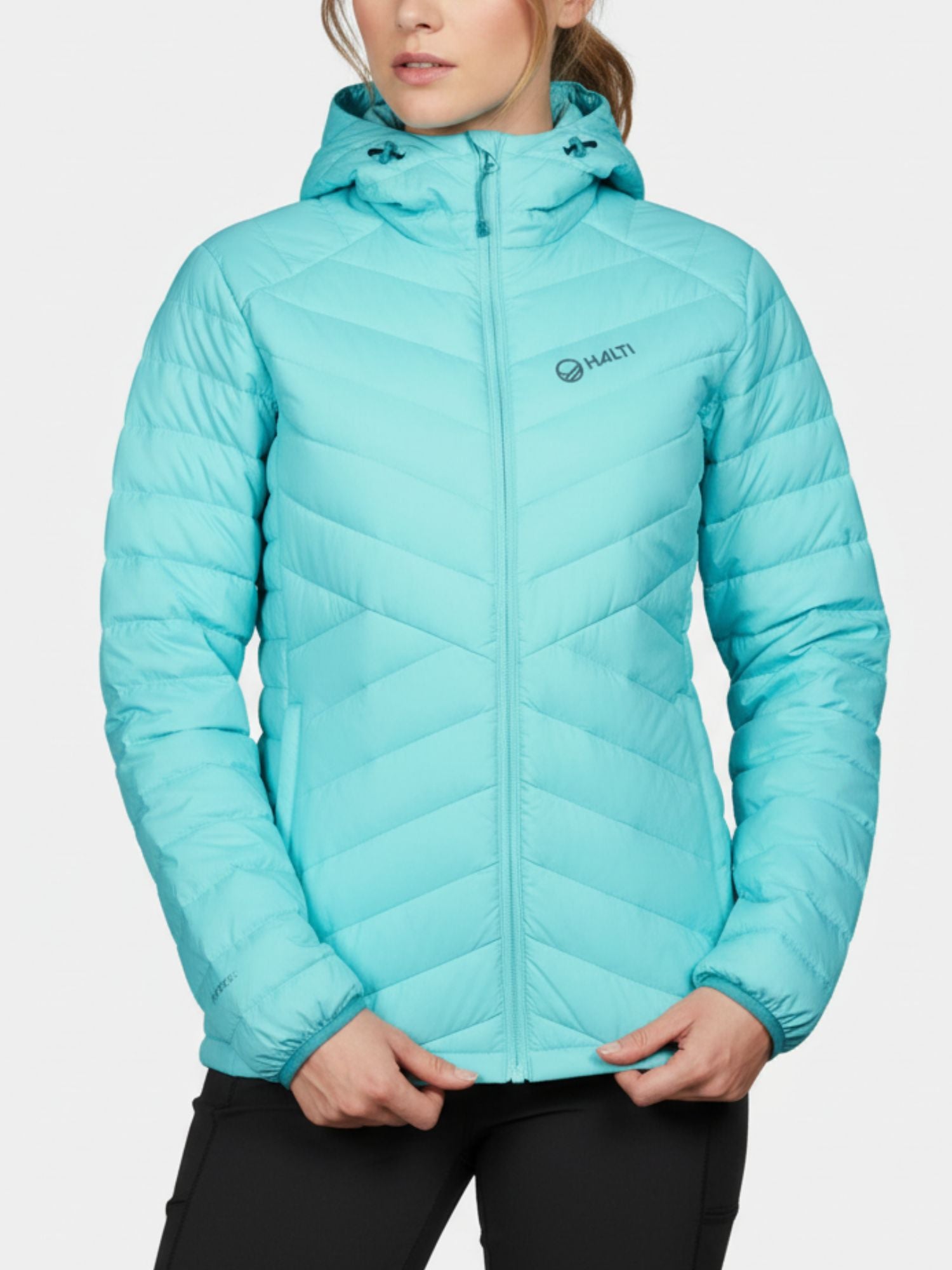 Naisella on yllään vaaleansininen Halti Huippu Untuvatakki, joka on valmistettu Pertex Quantumista, ja mustat housut yksiväristä taustaa vasten.