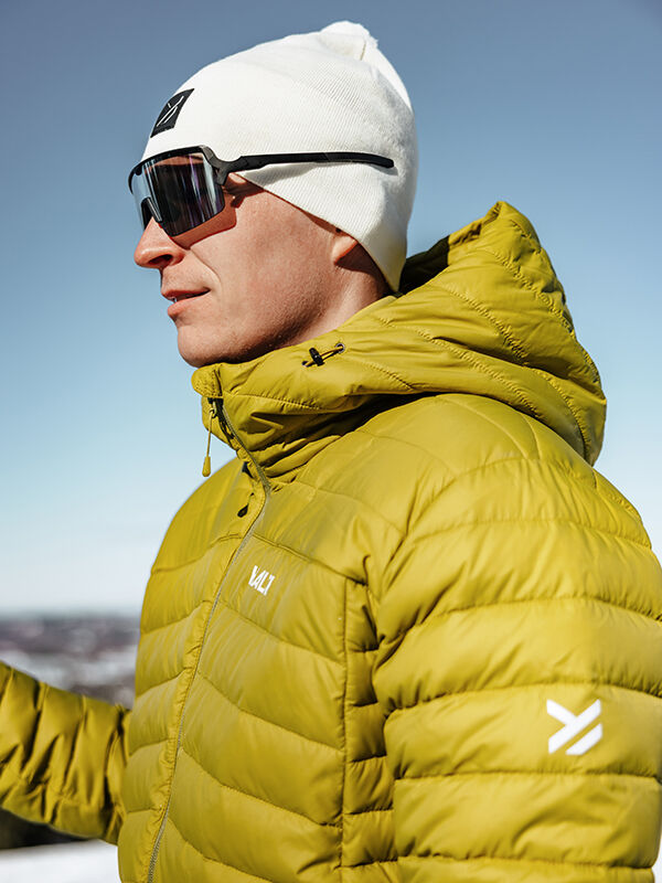 Mies keltaisessa Halti Huippu Untuvatakki Miesten, valkoinen pipo ja aurinkolasit seisoo ulkona aurinkoisena päivänä.