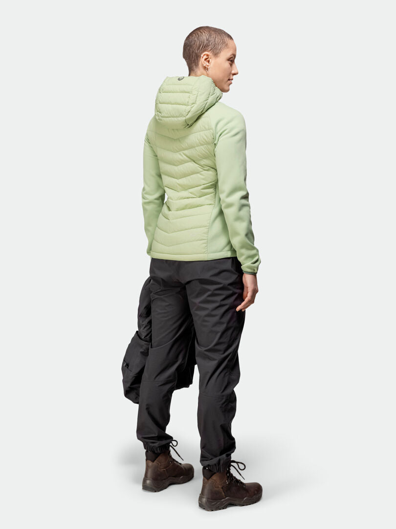 Henkilö on kasvot poispäin Halti Huippu Hybrid Untuvatakki Naisten takki, mustat housut, ruskeat saappaat, kädessä musta esine.