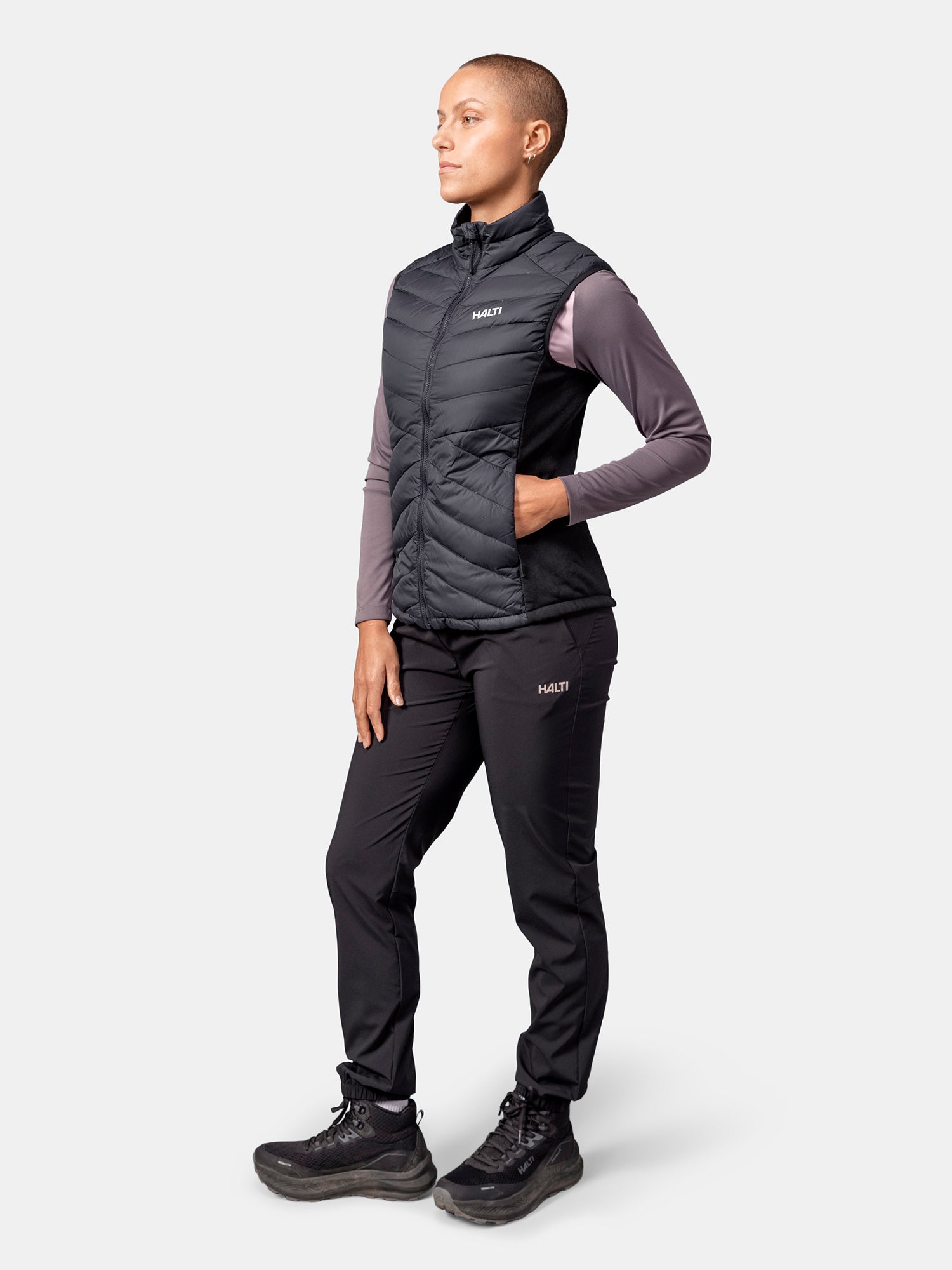 Henkilöllä on yllään musta Halti Huippu Hybrid Untuvaliivi -liivi, violetti pitkähihainen paita, mustat housut ja saappaat.