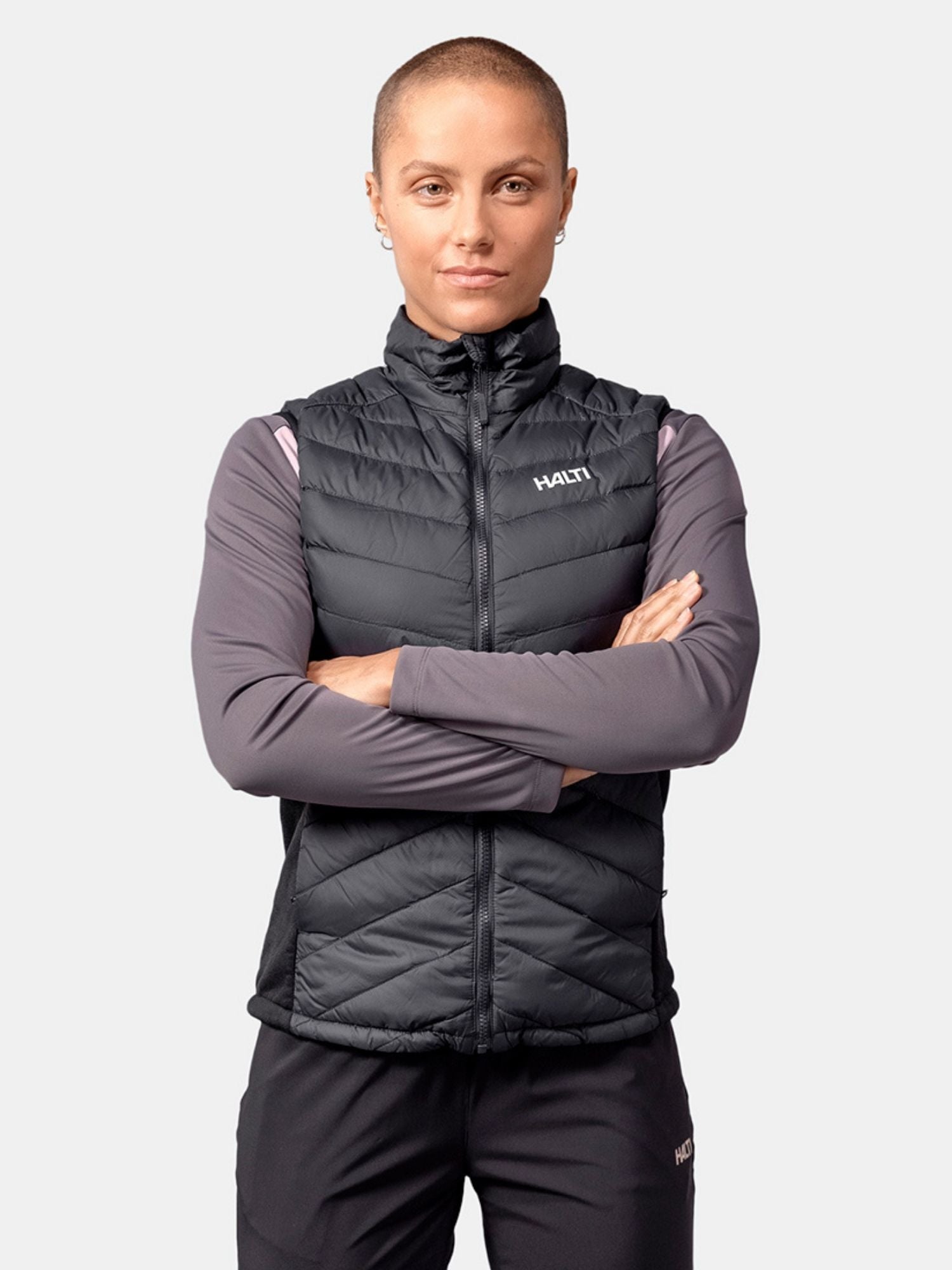 Tupeerattu henkilö pukeutuu Halti Huippu Hybrid Untuvaliivi Naisten pitkähihaisen paidan päälle, kädet ristissä.