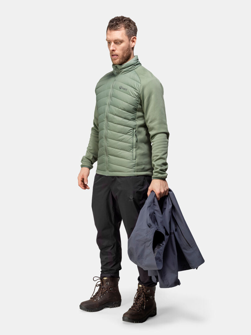 Vihreään Halti Huippu Hybridi Untuvatakki Miesten ja saappaisiin pukeutunut mies pitelee tummaa takkia tavallista taustaa vasten.