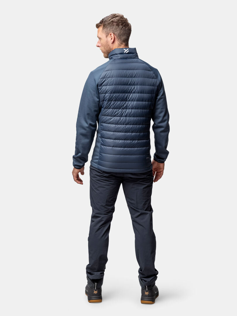 Mies seisoo kasvot menosuuntaan laivastonsinisessä Halti Huippu Hybrid Untuvatakki -miehessä, tummissa housuissa ja mustissa kengissä.