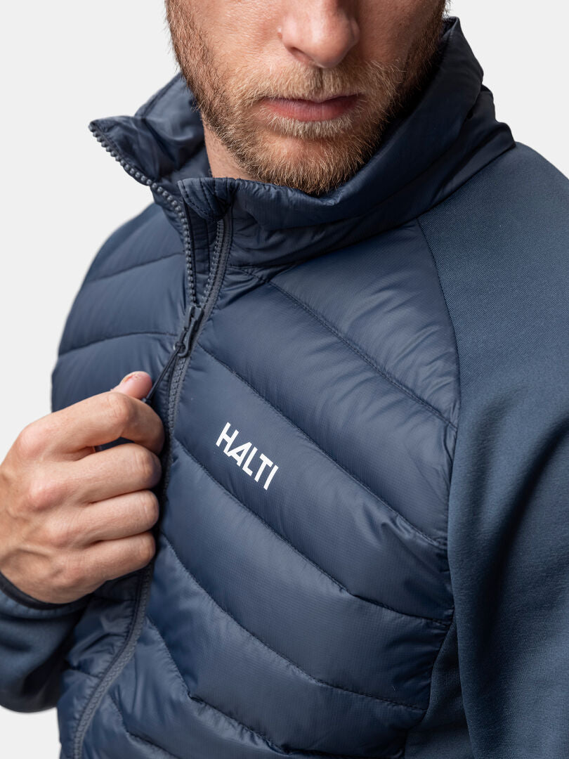 Mies vetää vetoketjun kiinni tummansiniseen Halti Huippu Hybridi Untuvatakki Miesten, joka on kuvattu rinta ylöspäin vaaleaa taustaa vasten.