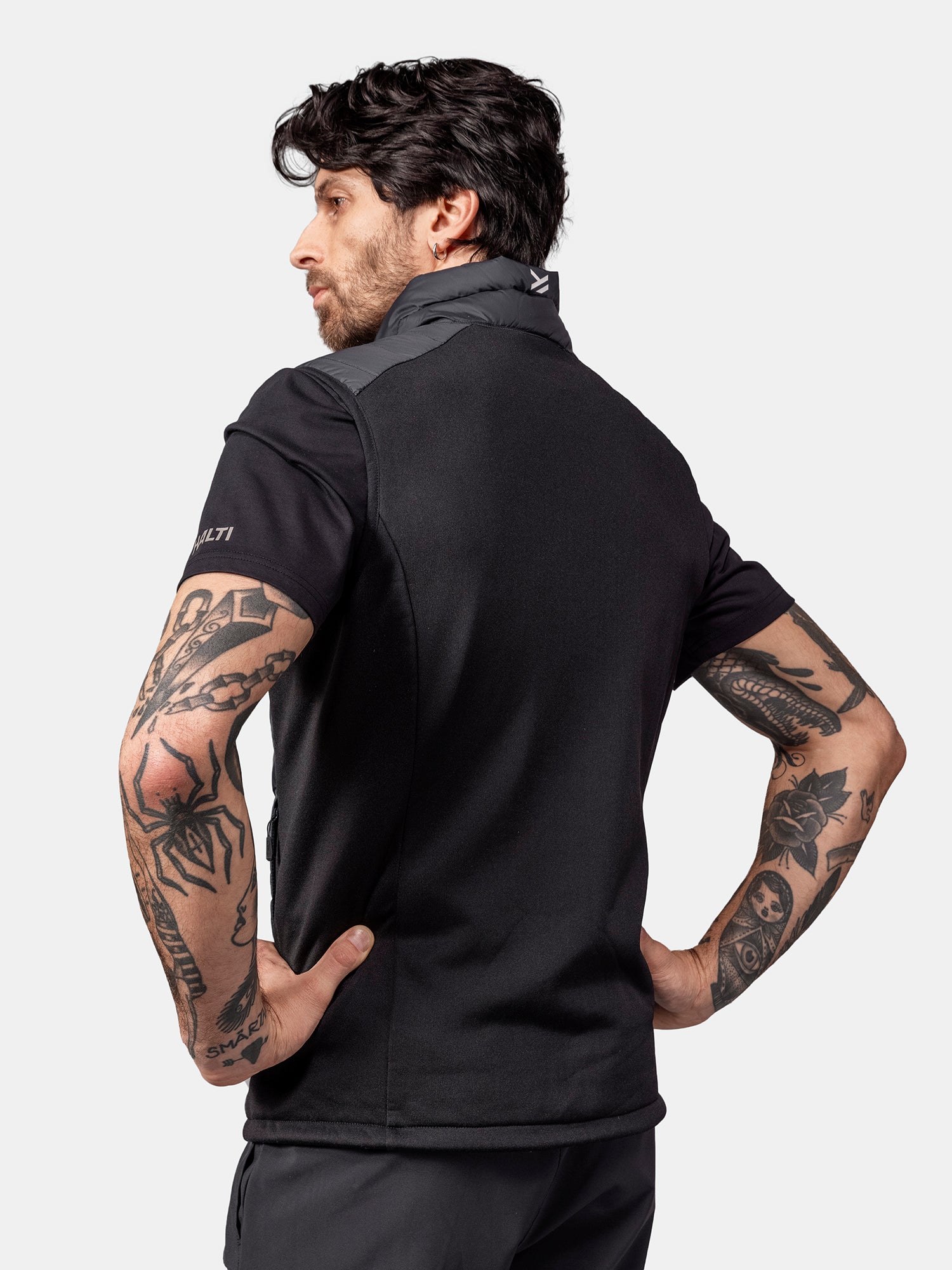 Tatuoidulla miehellä on yllään musta Halti Huippu Hybrid Untuvaliivi Miesten liivi, kasvot poispäin kädet lanteilla.