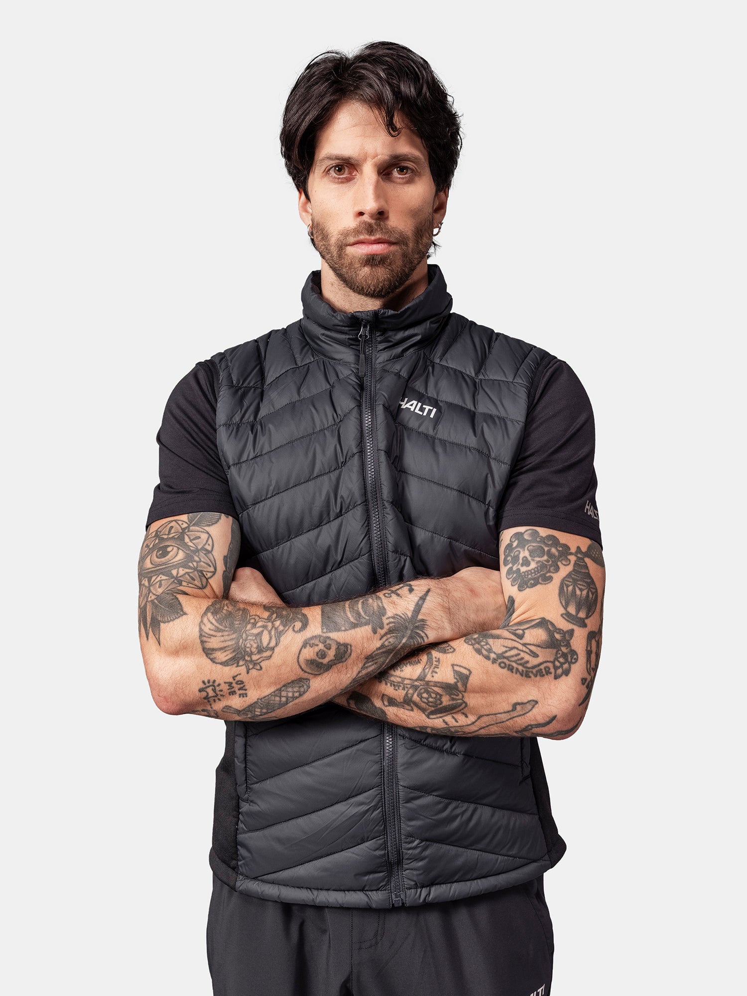 Tummahiuksinen ja tatuoitu mies pukeutuu mustaan Halti Huippu Hybrid Untuvaliivi Miesten mustan T-paidan päälle, kädet ristissä.