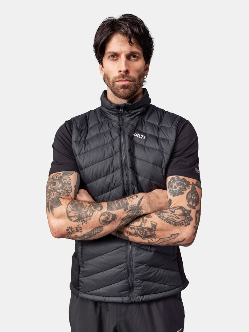 Tatuoitu mies mustassa Halti Huippu Untuvaliivi Miesten liivissä ja lyhythihaisessa paidassa, kädet ristissä.