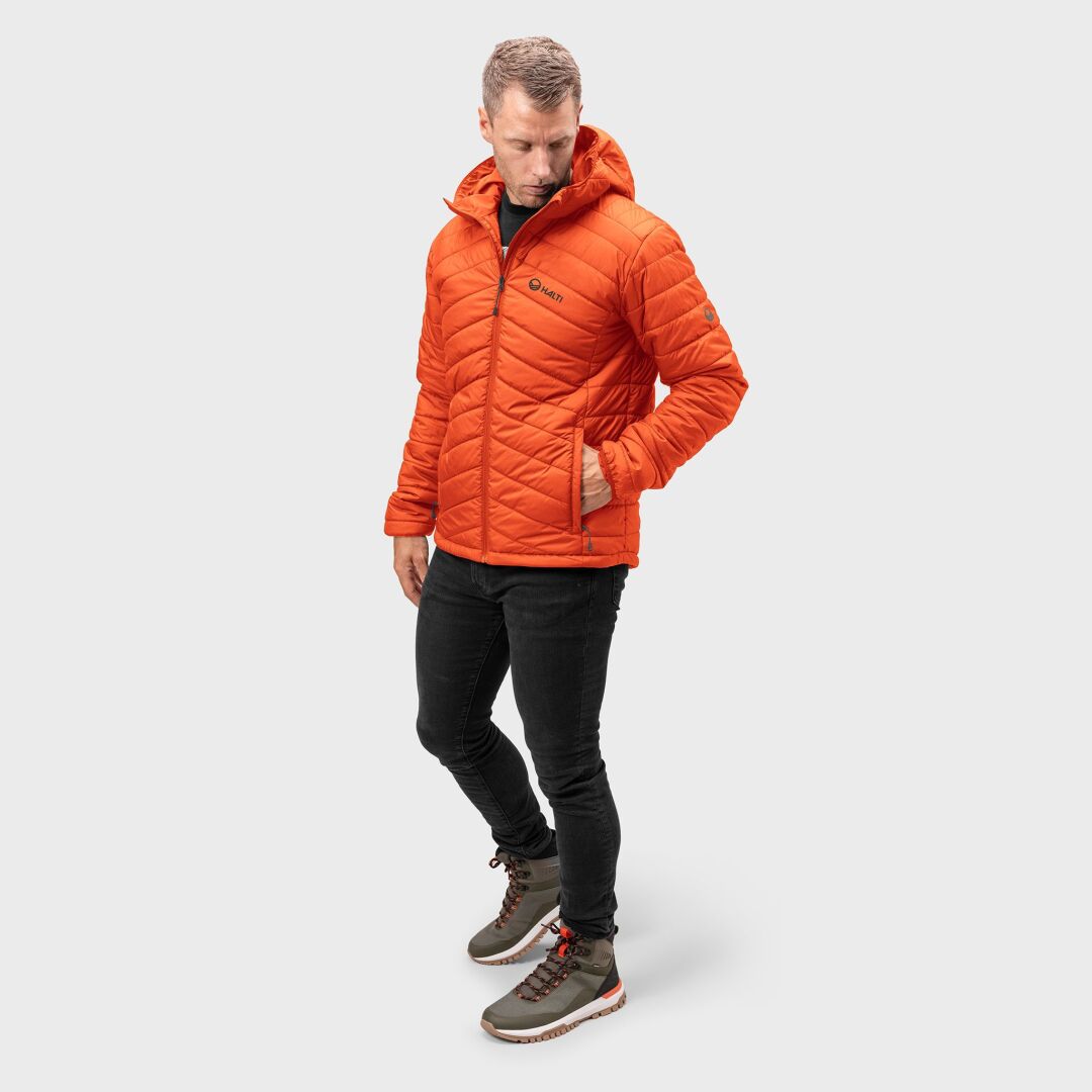 Mies yllään oranssi Halti Komu Kevytvanutakki Miesten -puku, mustat housut ja harmaat saappaat valkoista taustaa vasten.