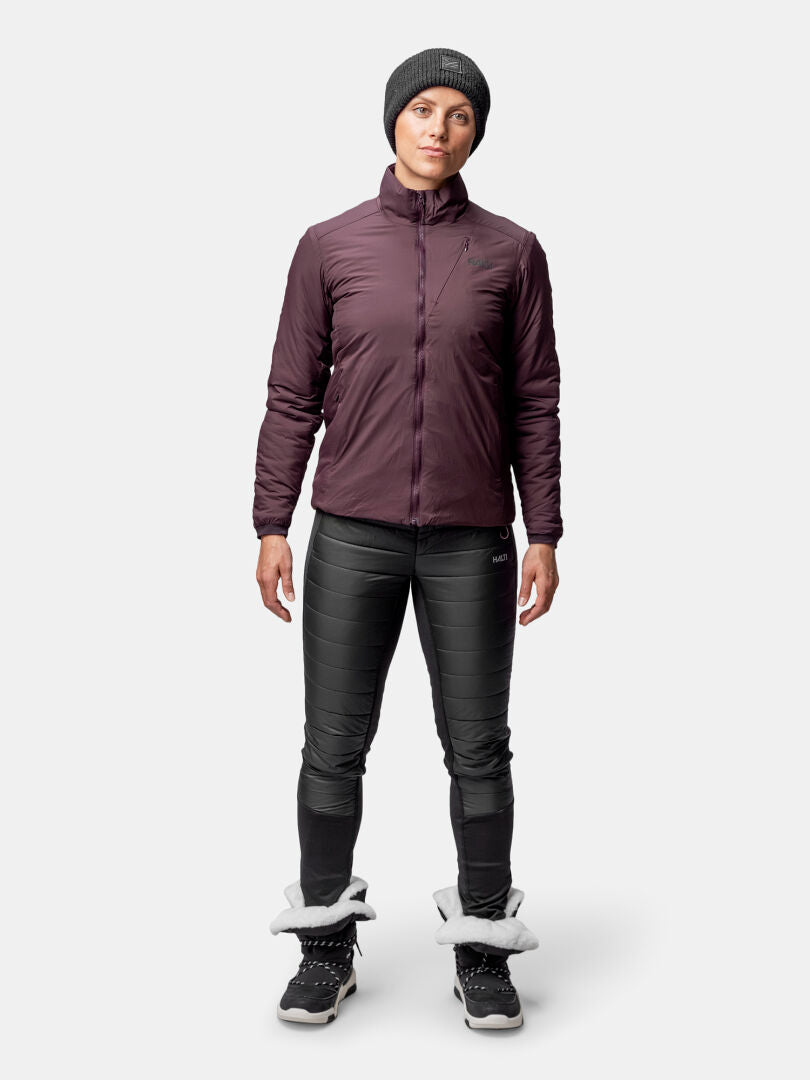 Naisella on ruskeankeltainen Halti Haave Primaloft Takki, mustat housut ja talvisaappaat valkoista taustaa vasten.