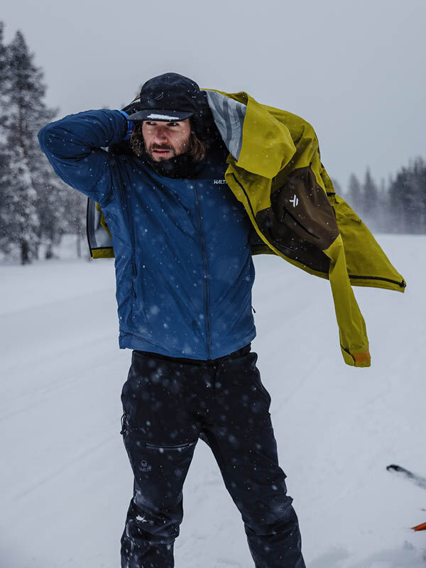 Talvivaatteisiin pukeutunut mies pukee keltaisen Halti Haave Primaloft Takki Miesten päälle seisoessaan ulkona putoavassa lumessa.