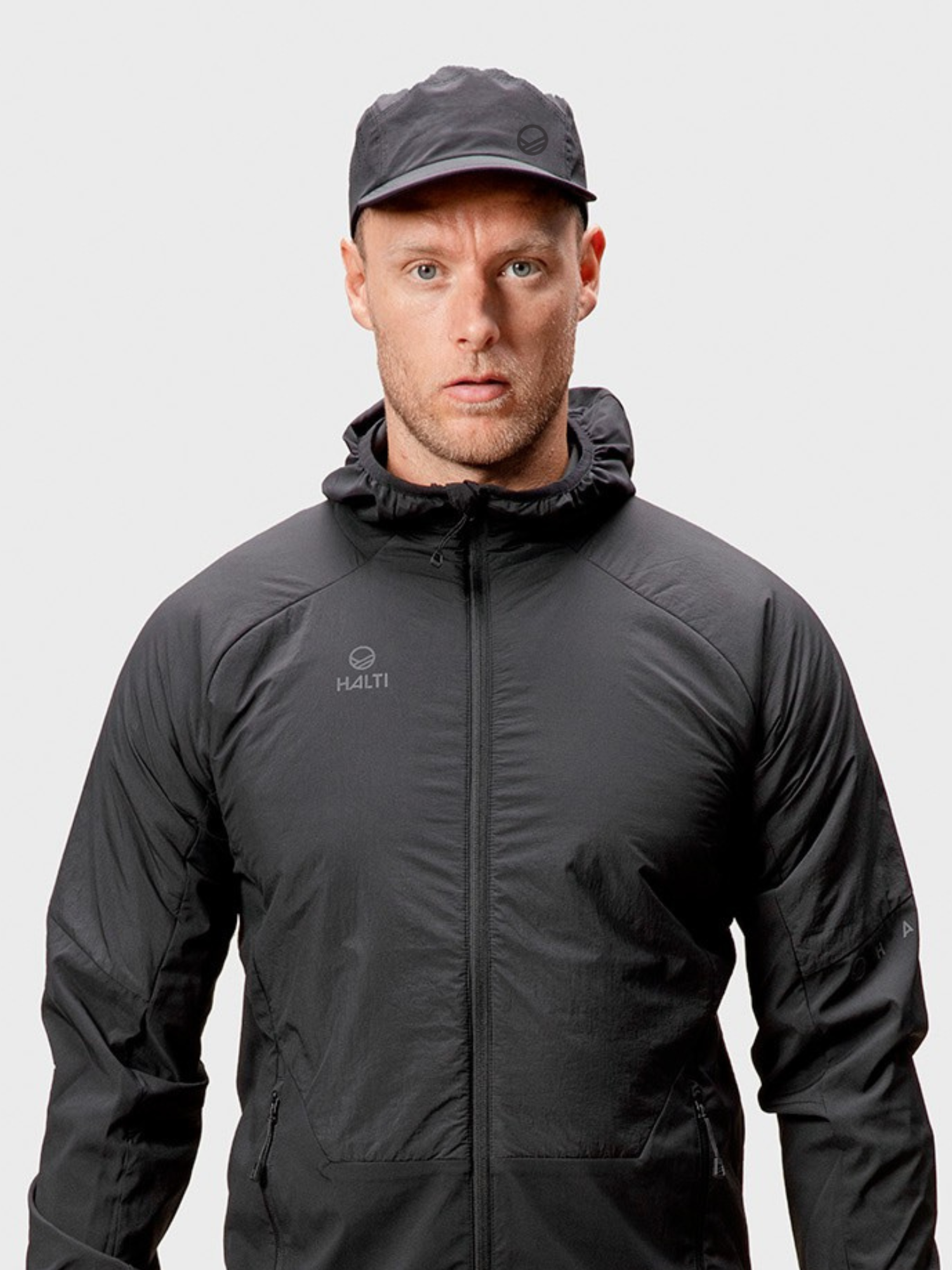Mustaan X-Stretch Rough -kangastakkiin ja Halti Aero II Lippis -takkiin pukeutunut mies seisoo vaaleaa, tavallista taustaa vasten.