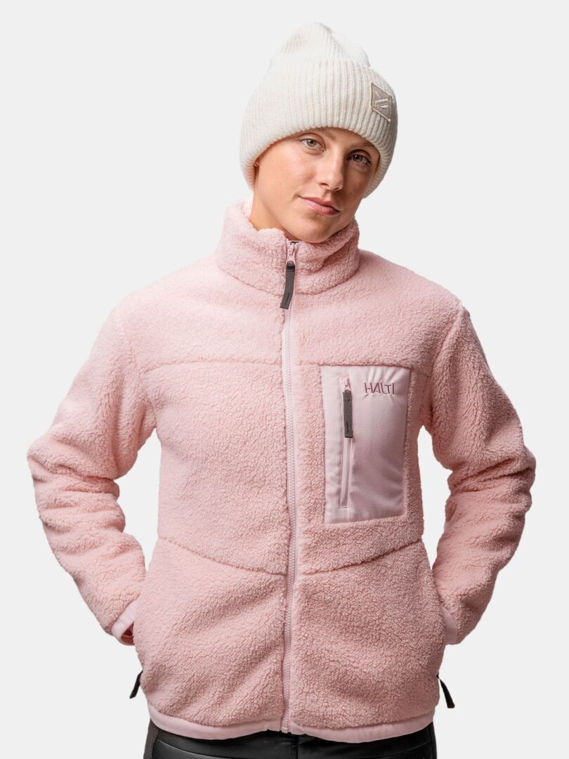 Henkilö, jolla on vaaleanpunainen fleecetakki ja Halti Huurre Pipo, kädet taskuissa, tavallista taustaa vasten.