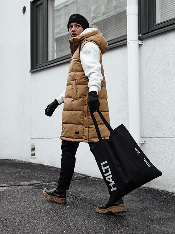 Henkilö, jolla on ruskehtava puskuriliivi, valkoinen huppari ja Viiri Fleece Hanskat, kävelee jalkakäytävällä mustan Halti-kangaskassin kanssa.