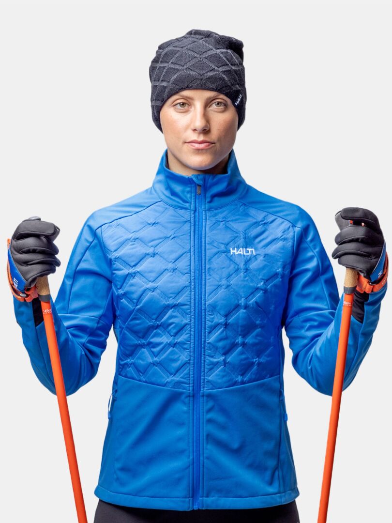 Stormwall-takkiin ja Halti Crostie Merinopipoon pukeutunut henkilö pitelee oransseja hiihtosauvoja valkoista taustaa vasten.