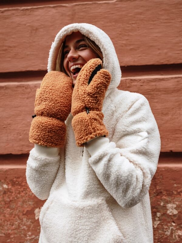 Henkilö, jolla on valkoinen hupullinen fleecetakki ja Halti Lykky Fleecehanskat, hymyilee kuvioidun ruskean seinän äärellä.