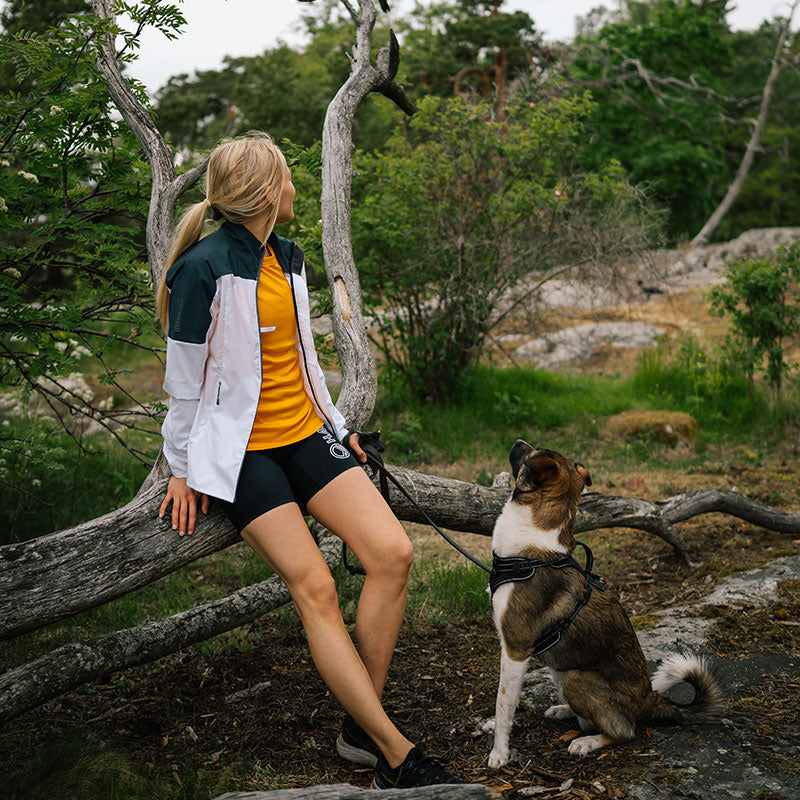 Nainen Halti Urbanite Naisten Treenitakki -takissa istuu tukilla koiransa kytkettynä metsässä.