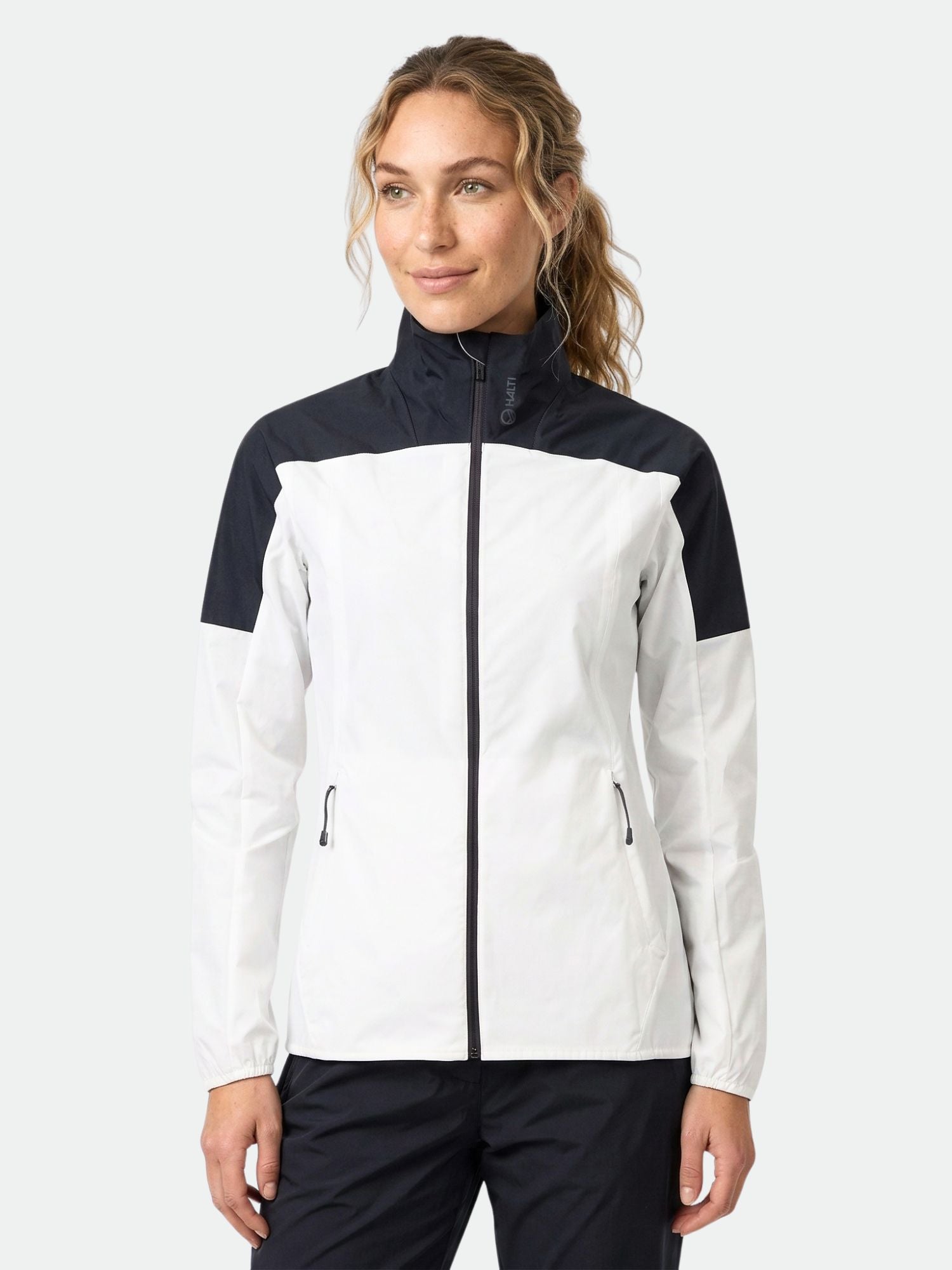 Naisella on yllään Halti Urbanite Naisten Treenitakki, mustavalkoinen pitkähihainen urheilutakki, tavallista taustaa vasten.