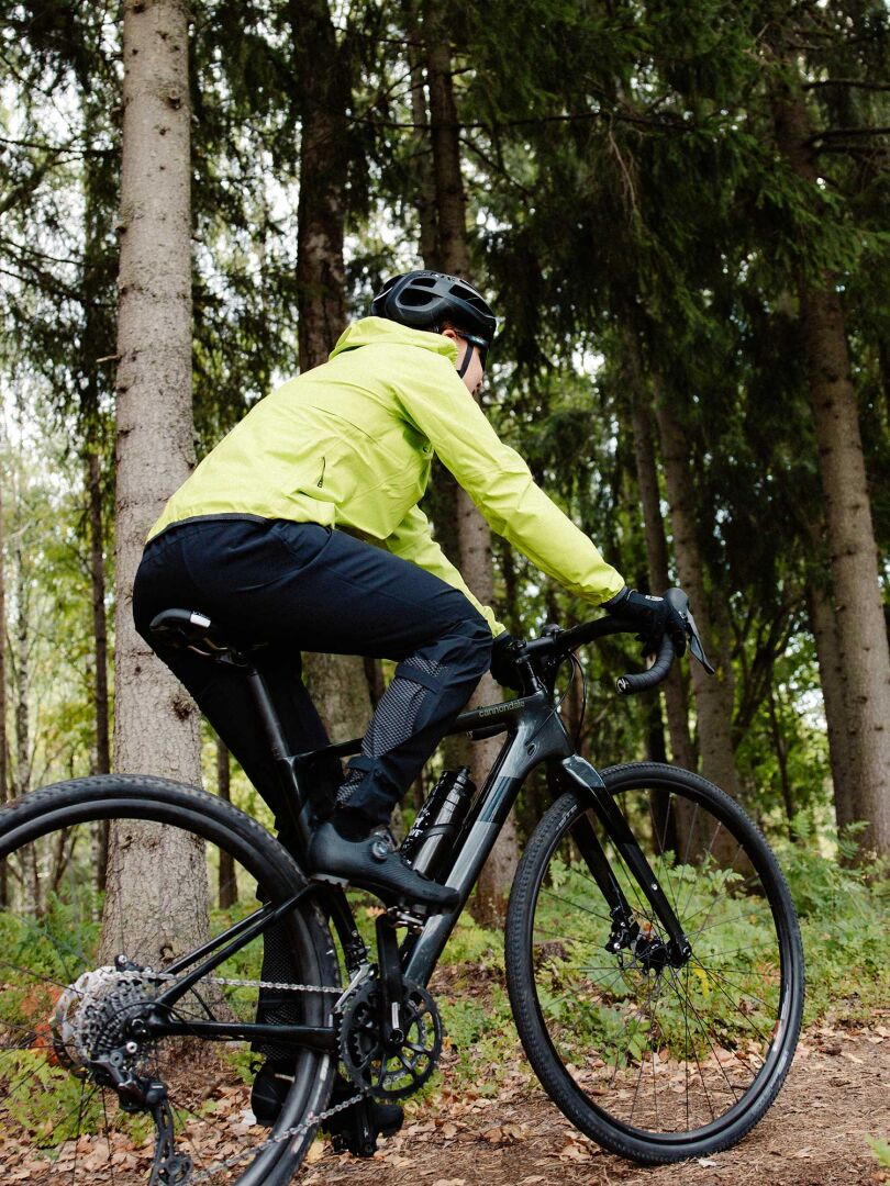 Henkilö keltaisessa Halti Liike Naisten 3L DrymaxX urheilutakissa ajaa mustalla pyörällä metsän hiekkapolulla.