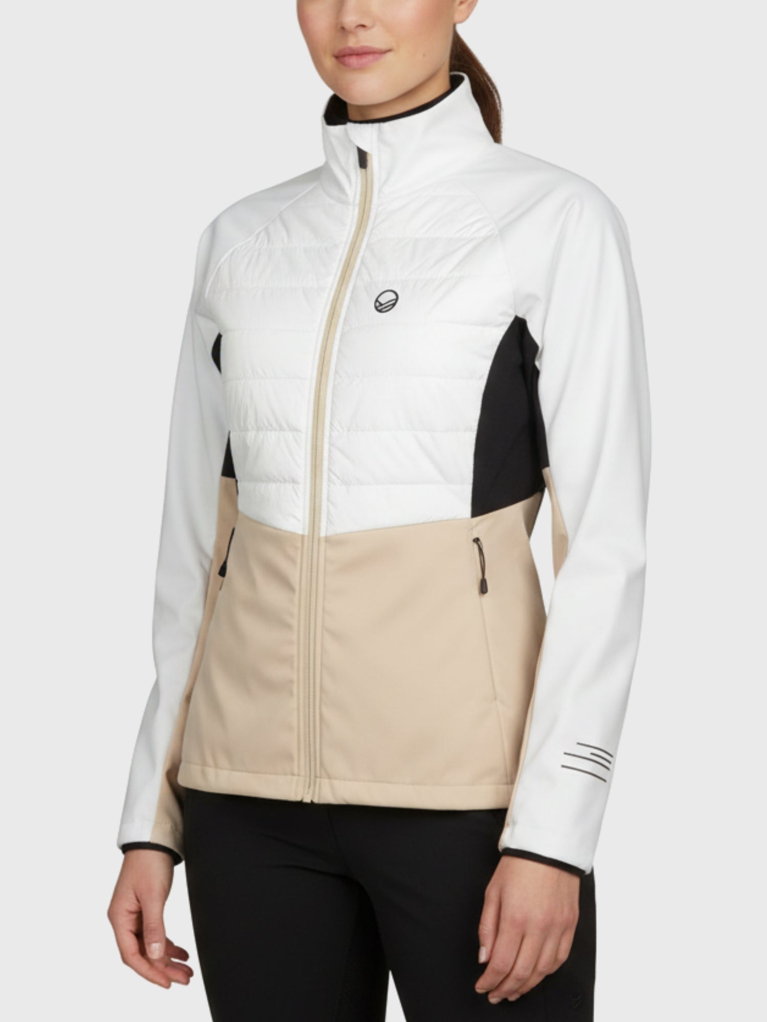 Naisella on yllään Halti Vinha Hybrid Hiihtotakki Naisten, jossa on valkoiset, beiget ja mustat paneelit, vetoketju edessä ja taskut.