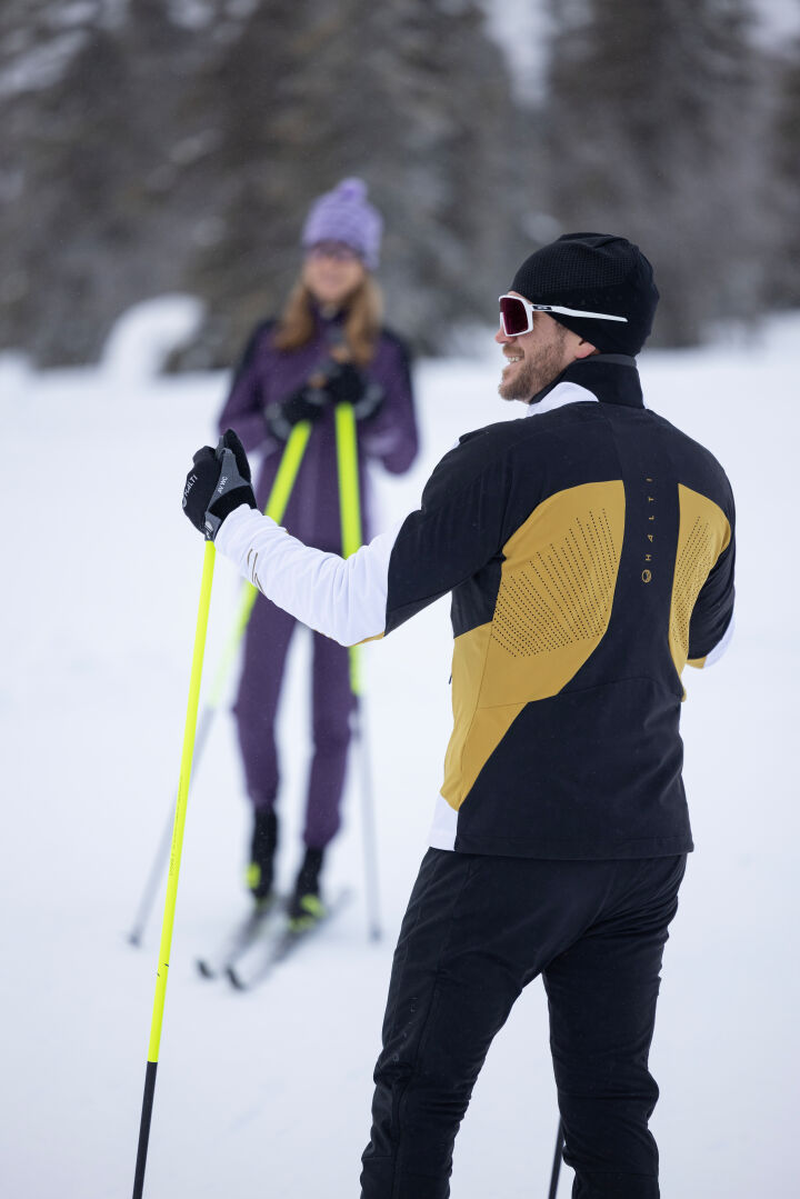 Kaksi henkilöä hiihtää Halti Veloce Hiihtotakki Miestenissä, molemmat pitävät sauvat lumisessa Stormwall® stretch-materiaalissa.