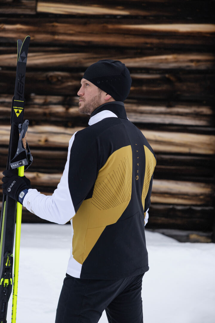 Mies Halti Veloce Hiihtotakki Miesten pitää suksia ja sauvoja kädessään lumen päällä puuseinän edessä.