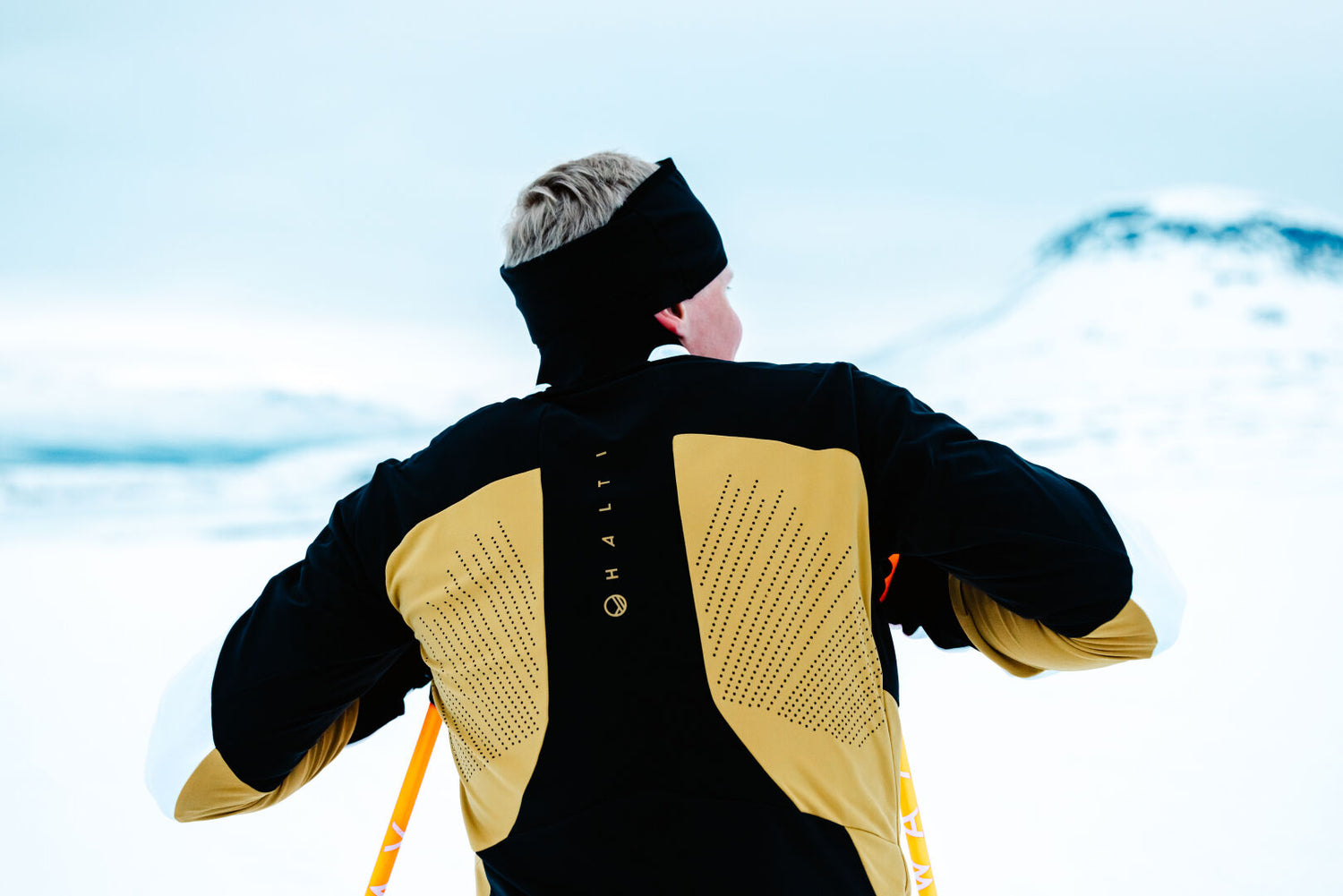 Henkilö Halti Veloce Hiihtotakki Miesten -hiihdossa seisoo lumimaisemassa kasvot poispäin hiihtosauvat kädessään.