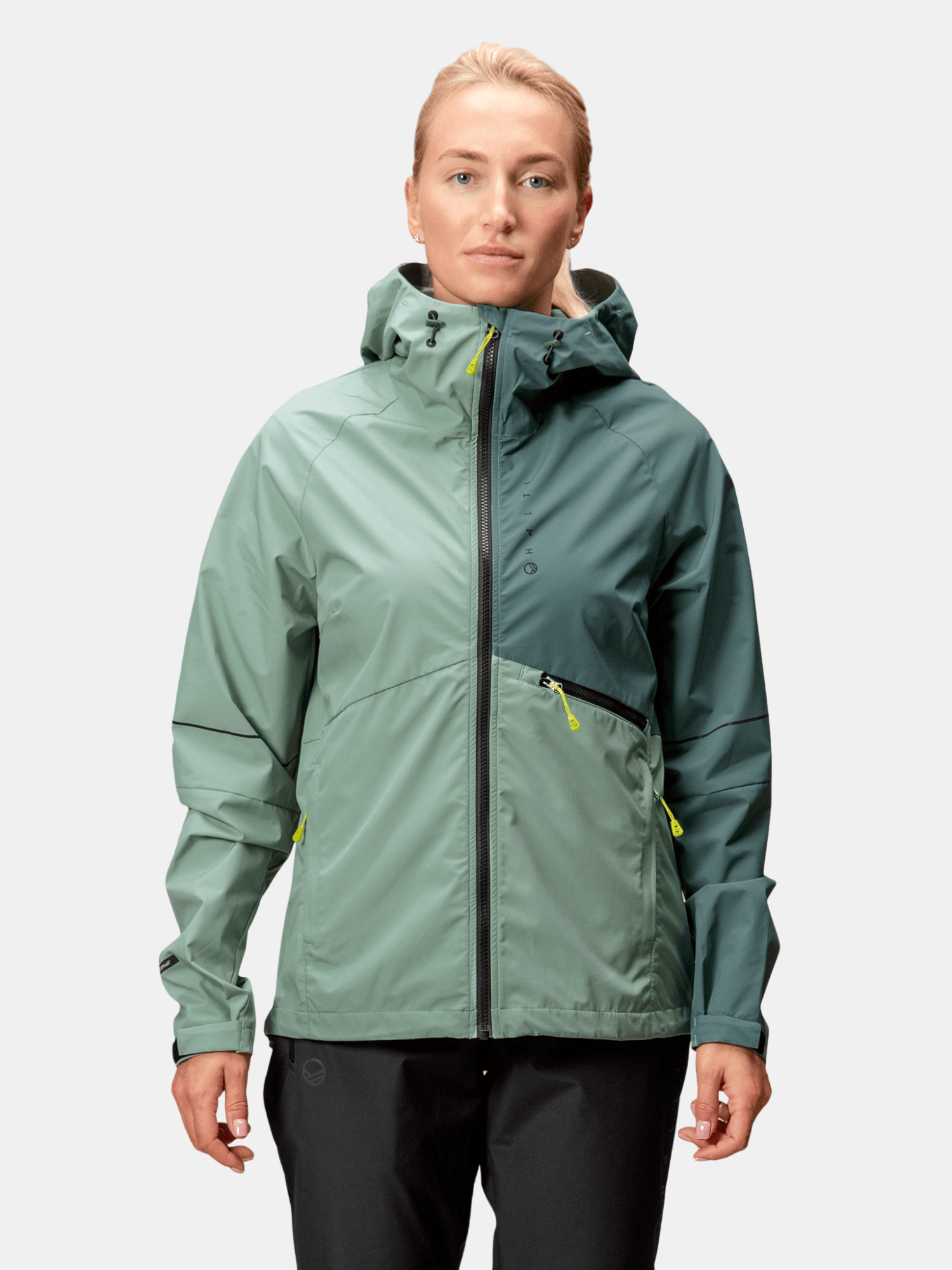 Nainen seisoo kasvot eteenpäin pukeutuneena Halti Shelter Stormwall 3L Takki Naisten -takissa ja mustissa housuissa.