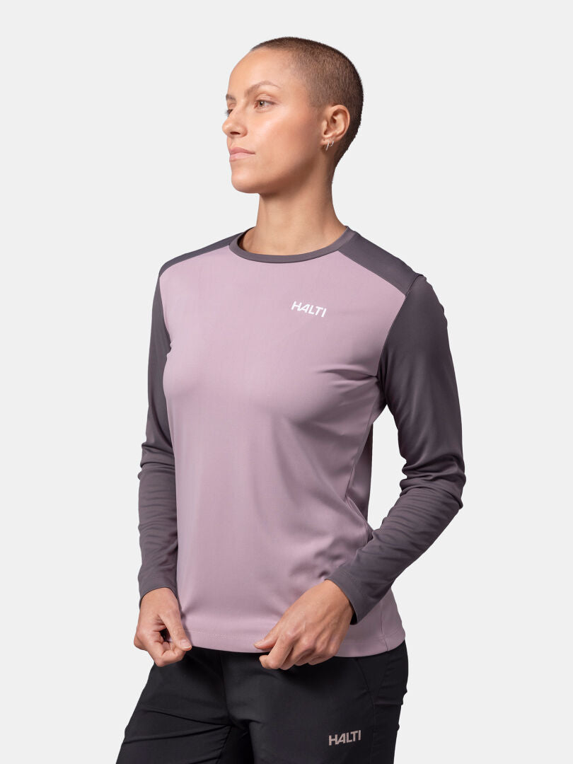 Nainen Halti Salves Treenipaita Naisten-pitkähihaisessa, vaaleanpunaisessa ja harmaassa naistenpuvussa seisoo tavallista taustaa vasten.