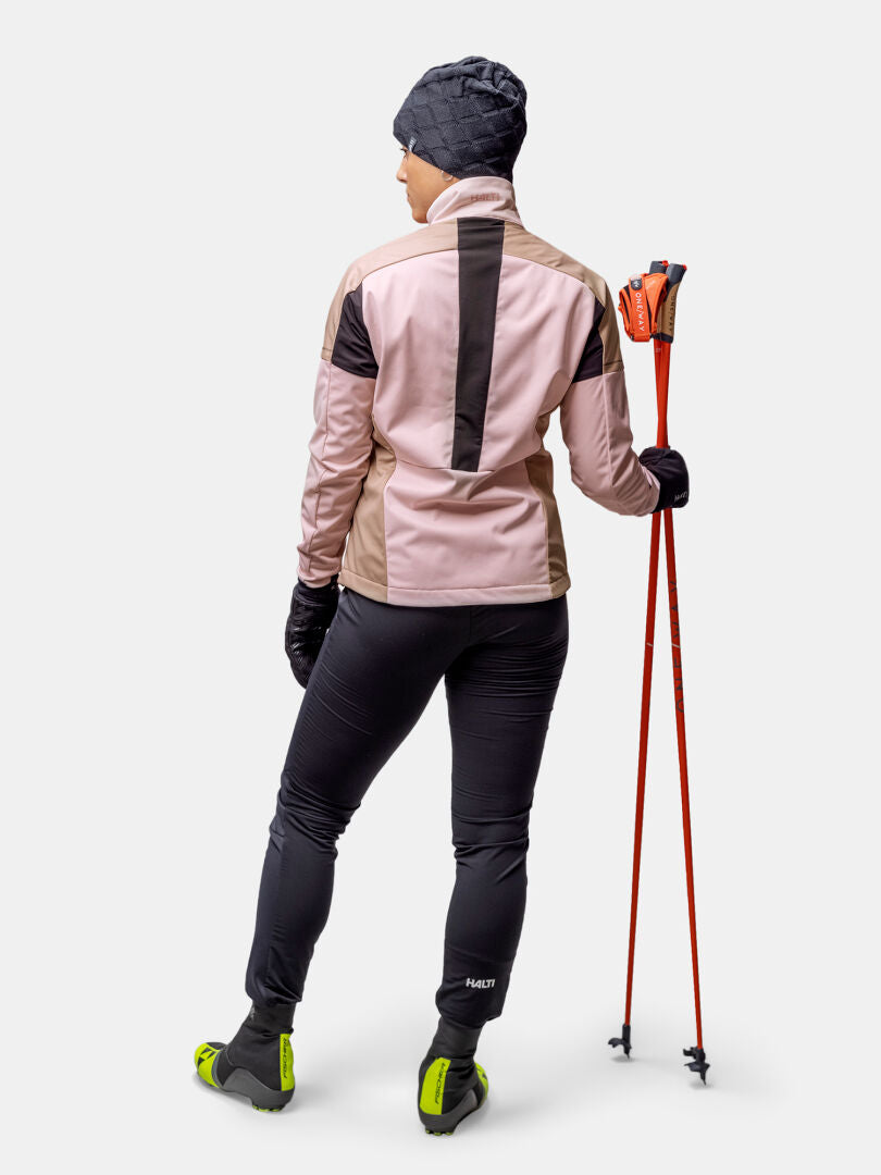 Henkilö Halti Huuru Maastohiihtopuku Naisten, punaiset hiihtosauvat kädessään, katsoo poispäin, valmiina maastohiihtoon.