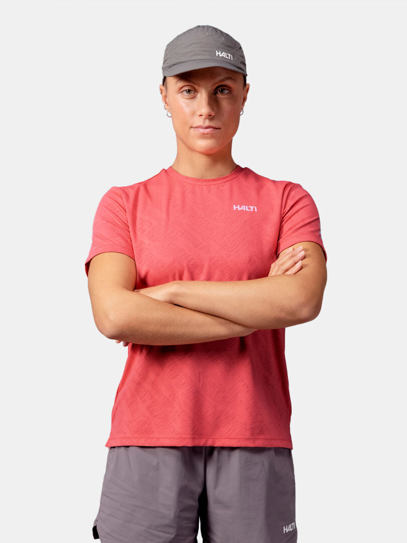Henkilö, jolla on vaaleanpunainen Halti Oulanka T-paita Naisten, harmaat shortsit ja lippis, seisoo kädet ristissä tavallista taustaa vasten.