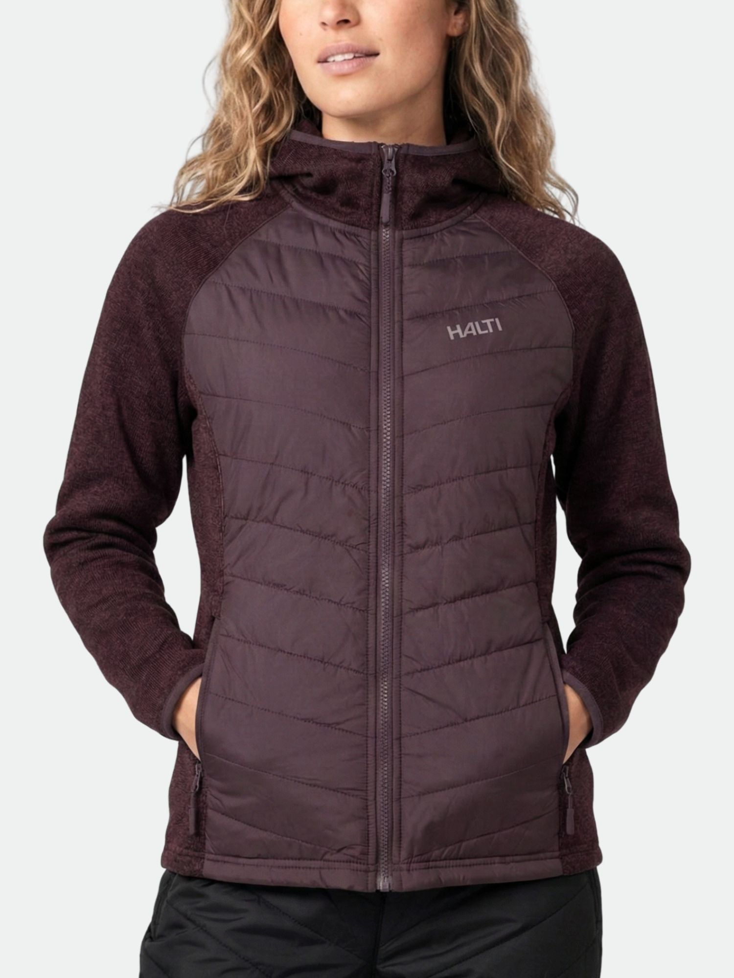 Henkilö, jolla on yllään Halti Streams Hybrid Välitakki Naisten ruskeankeltainen huppu ja kädet taskuissa, yksivärisellä taustalla.