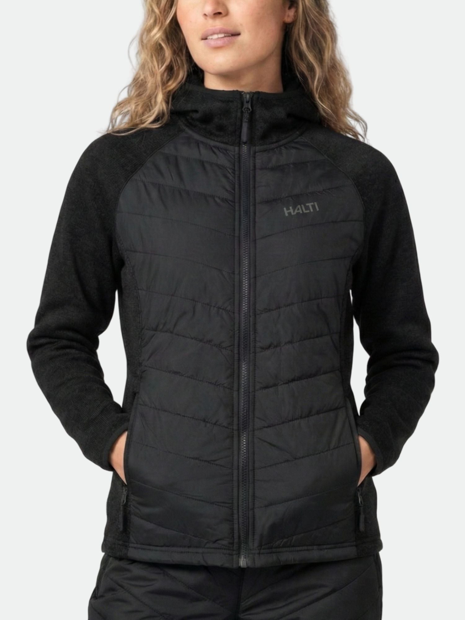Henkilöllä on yllään musta Halti Streams Hybrid Välitakki Naisten, kädet taskuissa, vaaleaa taustaa vasten.