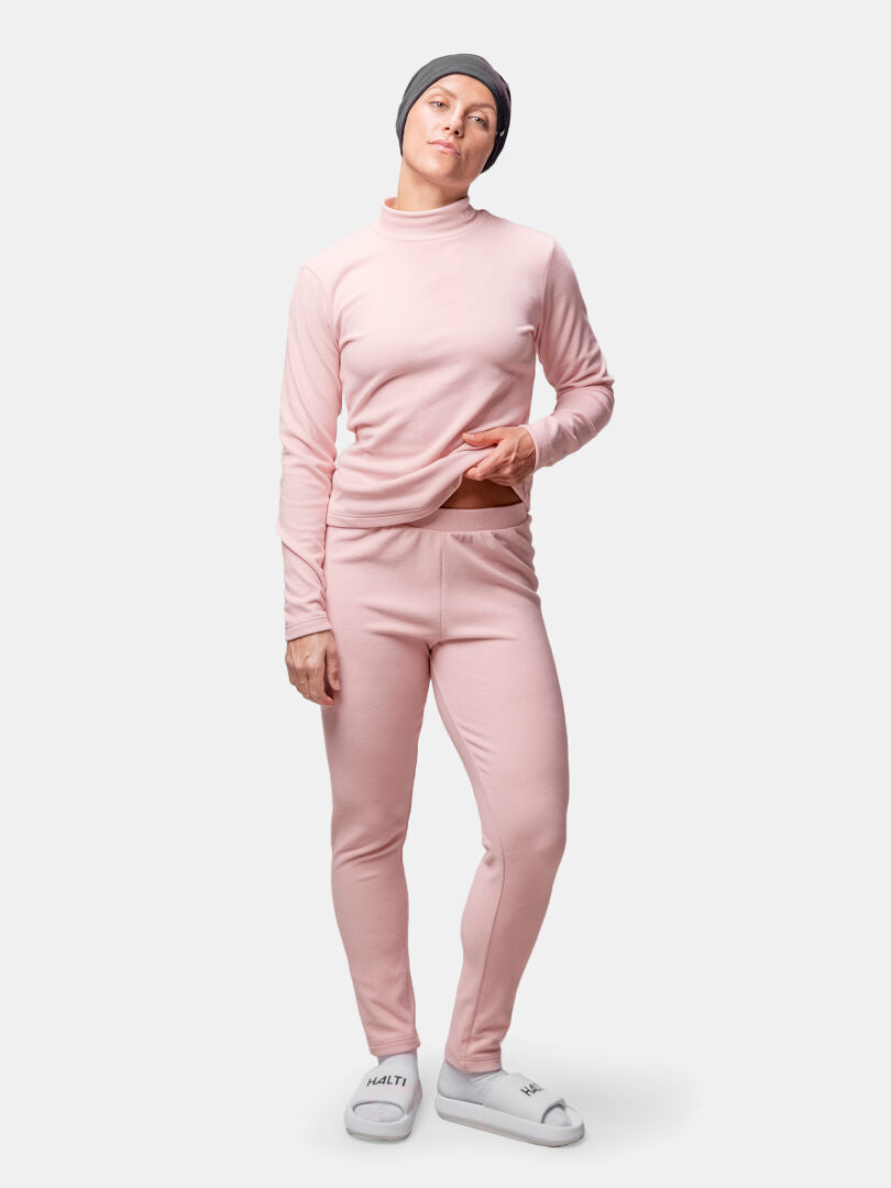 Henkilöllä on Halti Trone Fleece Paita Naisten ja housujen setti, harmaa pipo, valkoiset liukusukat, tavallista taustaa vasten.