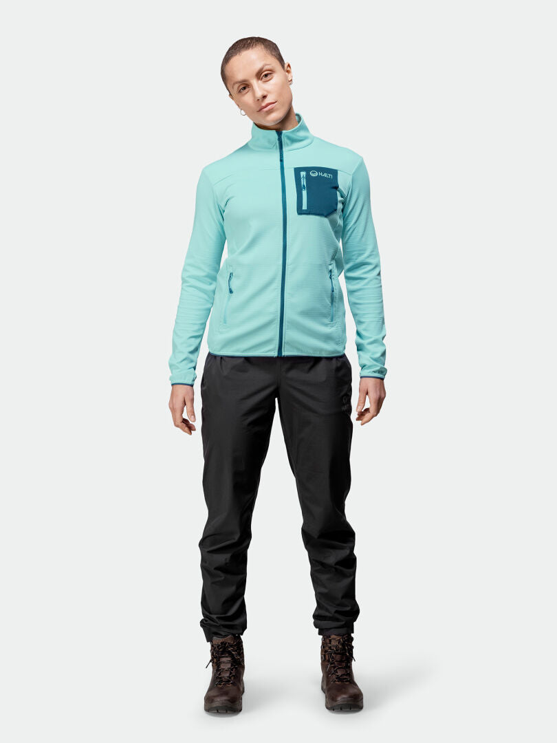 Henkilöllä on yllään vaaleansininen Halti Toivo Välitakki Naisten paita, mustat housut ja ruskeat vaelluskengät yksiväristä taustaa vasten.