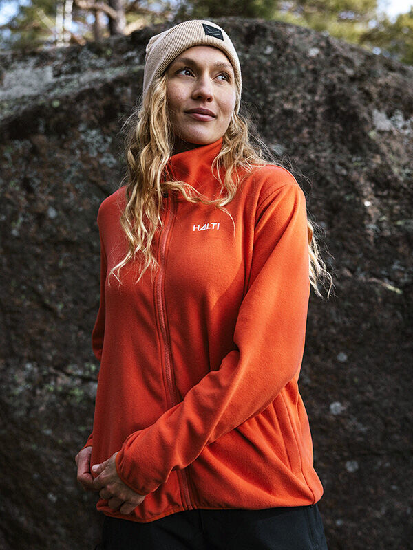 Oranssiin Halti Rockmoon Fleecetakki Naisten ja beigeen pipoon pukeutunut nainen seisoo ulkona suuren kiven äärellä.