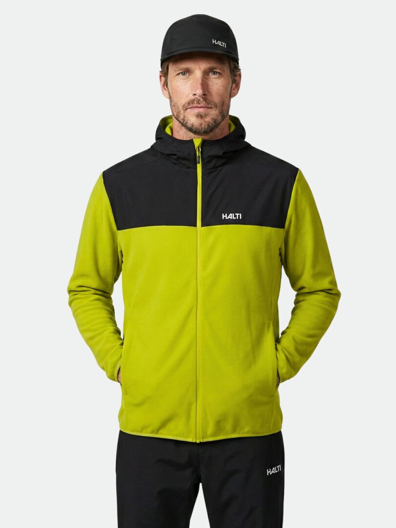 Mies, jolla on musta lippis ja Halti Rockmoon Fleecehuppari, seisoo kädet taskuissa tavallista taustaa vasten.